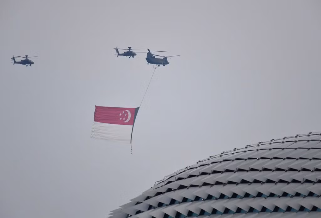 Cuộc duyệt binh của Singapore huy động nhiều loại khí tài thuộc hàng hiện đại nhất thế giới. Trong ảnh, đội hình trực thăng vận tải Chinook treo quốc kỳ Singapore bay qua quảng trường với sự hộ tống của biên đội trực thăng Apache. Nguồn ảnh: Mediacorp