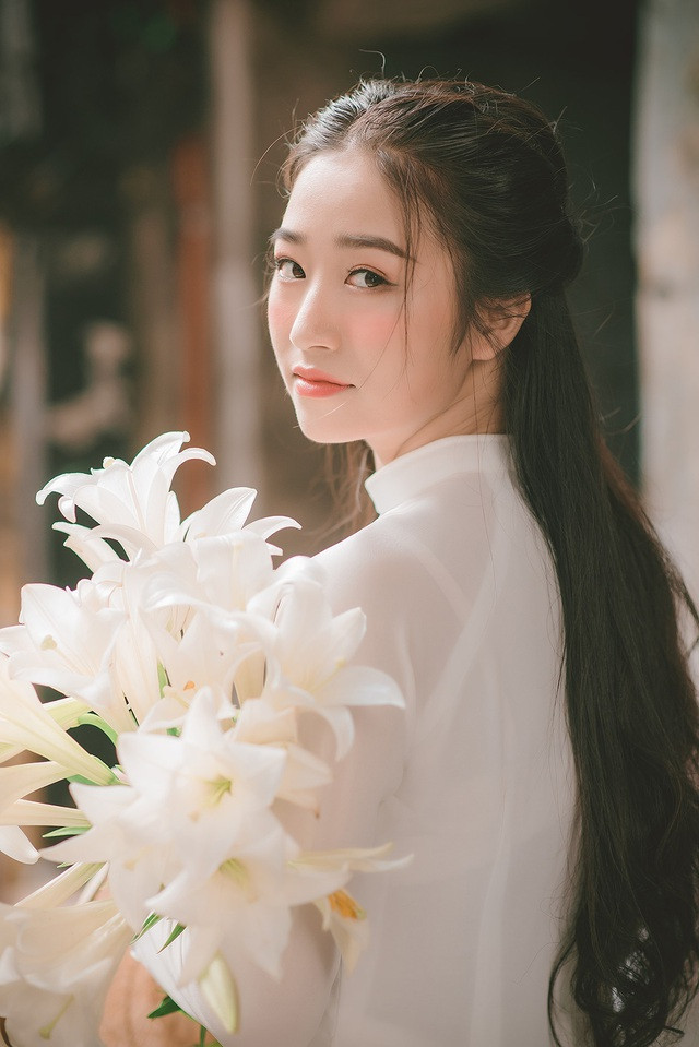 Trong tà áo dài trắng, Ngọc Mai trang điểm đơn giản, ôm bó hoa loa kèn vào lòng và nở nụ cười tươi.