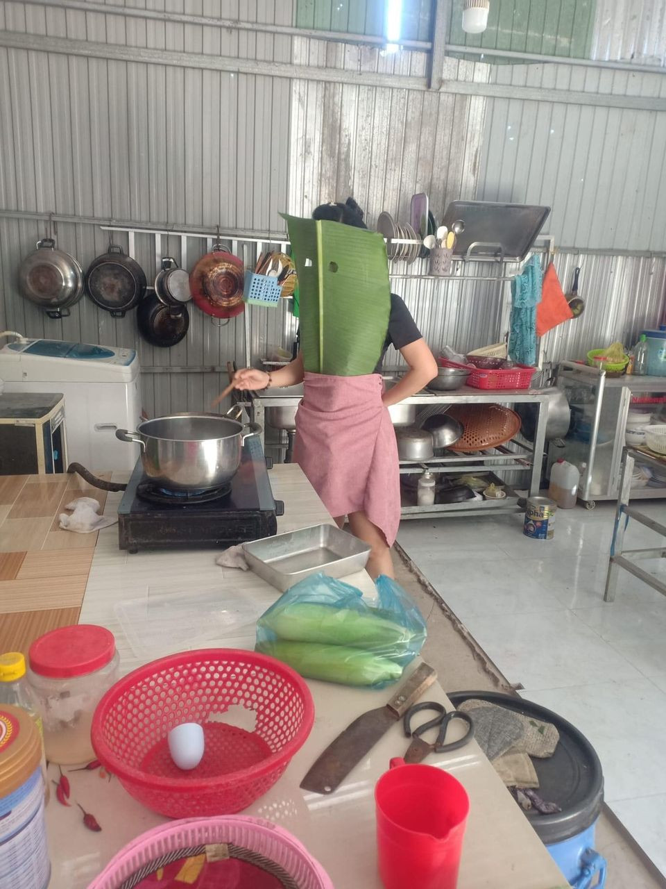 Để chuẩn bị cho màn rán cá không bị bắn của mình, người phụ nữ đã ra vườn, chọn chiếc lá chuối phù hợp sau đó đeo lên người và điều thú vị là không quên khoét 2 cái lỗ để nhìn chảo cá rán.