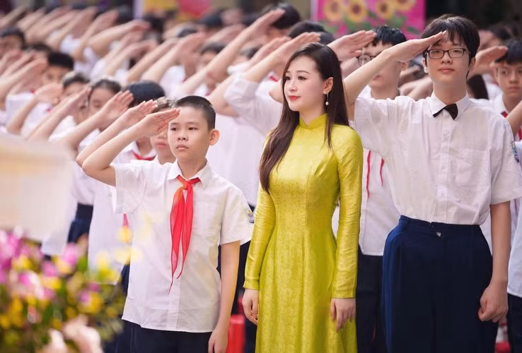 Đứng dưới sân trường, cô giáo có gương mặt thanh tú diện áo dài truyền thống màu vàng chanh nổi bật. Mái tóc dài thướt tha và lối trang điểm nhẹ nhàng càng tôn nên vẻ thanh lịch, dịu dàng.