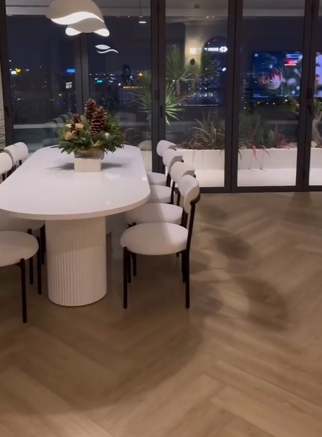 Phòng ăn có view hướng ra thành phố rực rỡ ánh đèn nhờ cửa kính lớn. Trên bàn ăn đặt chậu thông Noel, phía trên là đèn trần nghệ thuật. Ảnh chụp màn hình