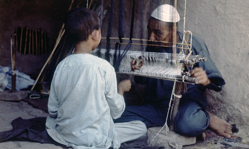 Người đàn ông lớn tuổi chỉ dạy cho cậu bé cách sử dụng khung cửi.