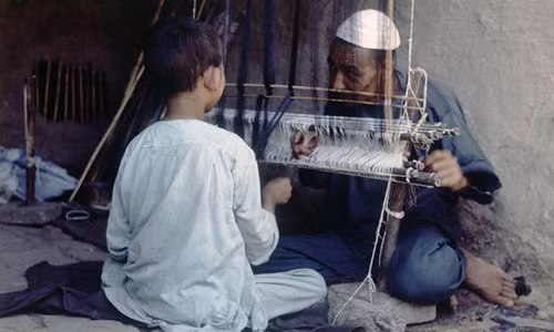 Người đàn ông lớn tuổi chỉ dạy cho cậu bé cách sử dụng khung cửi.