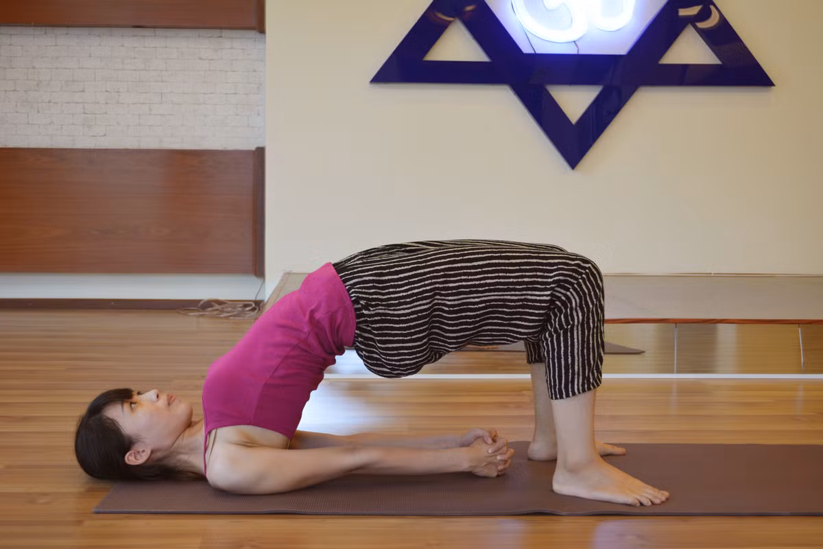 Tư thế cây cầu. Nằm ngửa,đầu gối co lên. Hai bàn chân cách nhau 1 khoảng bằng hông (khoảng cách giữa hông và gót chân bằng 1 bàn tay. Hai cánh tay để dọc theo thân người, lòng bàn tay úp xuống. Hoi thu cằm xuống ngực để thư giãn gáy. Hít vào, thở ra, đầy bàn chân xuống sàn để nâng hông và lưng lên khỏi sàn cao hết mức có thể. Giữ hai bàn chân nằm nguyên dưới đầu gối, đặc biệt nếu các bạn có vấn đề về khớp gồi. Ảnh: Vyogaworld.