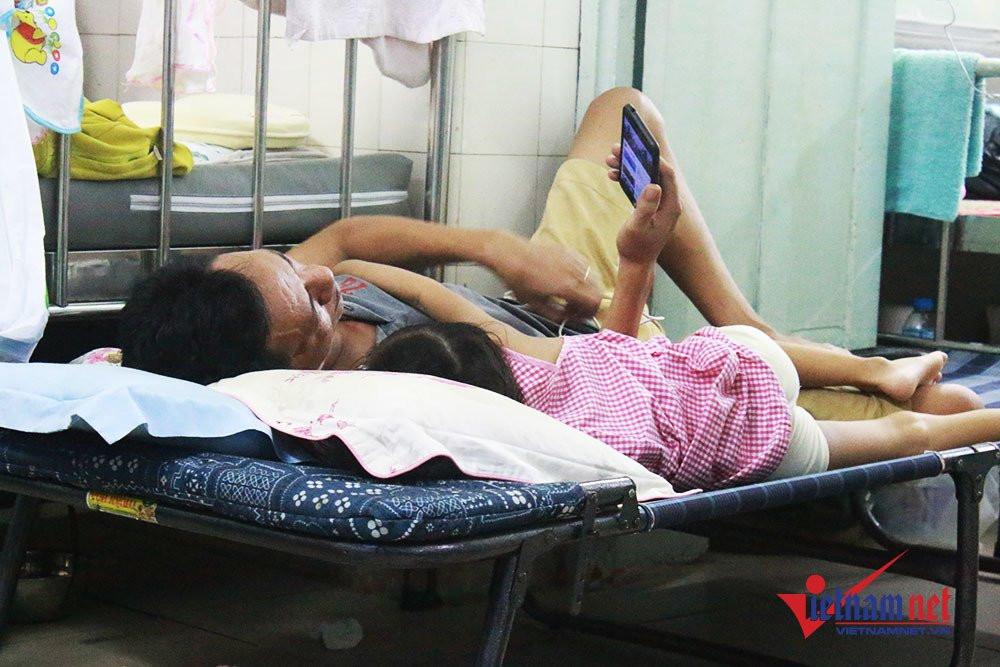 Cho con xem hoạt hình - phút giải trí hiếm hoi của cha cùng con gái trong bệnh viện. Do thời tiết nắng nóng, bệnh nhi đông nên nhiều phụ huynh ra ngoài sân của bệnh viện ngồi. Đến gần 9h tối, họ quay trở vào để thay nhau chăm sóc con, cháu đang nằm tại khoa Nội Tổng hợp.