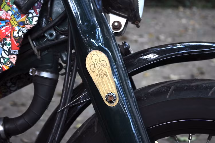 Để có thể lắp được bánh trước lớn cho xe, cặp phuộc giò gà đã phải nới rộng ra. Cụm đèn pha chóa tròn cùng đèn xi-nhan được chuyển xuống dưới dè trước. Hai bên "giò gà" của xe còn được chủ nhân trang trí bằng những miếng ốp bằng đồng CNC hình biểu tượng hoa bách hợp đầy vẻ cổ điển.