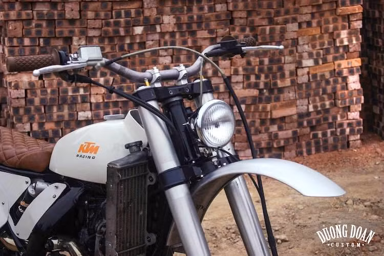 Lột bỏ toàn bộ dàn vỏ nhựa của KTM 350 EXC nguyên bản, DuongDoan đã bắt đầu từ "xác xe" gồm khung, máy, hệ thống phuộc gắp và 2 bánh xe. Ở phía trước, nằm giữa cặp phuộc USD hành trình lớn nguyên bản do WP sản xuất nay là một chiếc đèn pha tròn cổ điển, nhỏ gọn. Bên dưới dèn pha, xưởng độ đã gò lại cho 350 EXC chiếc chắn bùn bằng nhôm đúng "chất cào cào".