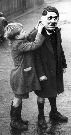 Một bé trai giúp bạn đeo mặt nạ có hình trùm phát xít Hitler. Ảnh chụp ở London năm 1938.
