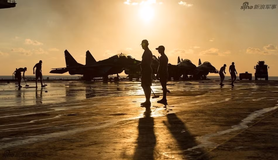 Những thủy thủ đang dọn dẹp sàn tàu dưới ánh hoàng hôn xế chiều, phía xa là những chiếc tiêm kích hạm Su-33 của Không quân Hải quân Nga nằm im đợi lệnh. Nguồn ảnh: Sina.