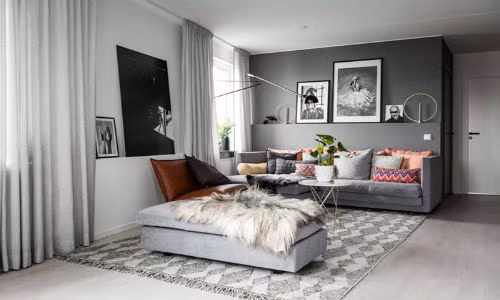 Không gian phòng khách được bố trí ngay cạnh cửa ra vào. Cách chọn lựa nội thất không có quá nhiều sự đặc biệt. Tuy nhiên, cách sắp xếp màu sắc lại là cả một sự tinh tế. Bức tường phía sau sofa được chọn lựa với màu xám đậm, giúp sofa có màu xám sáng trở thành điểm nhấn tinh tế cho không gian. Để cân bằng màu sắc, tấm thảm trải sàn với họa tiết quả trám màu xám được đặt ngay ngắn.