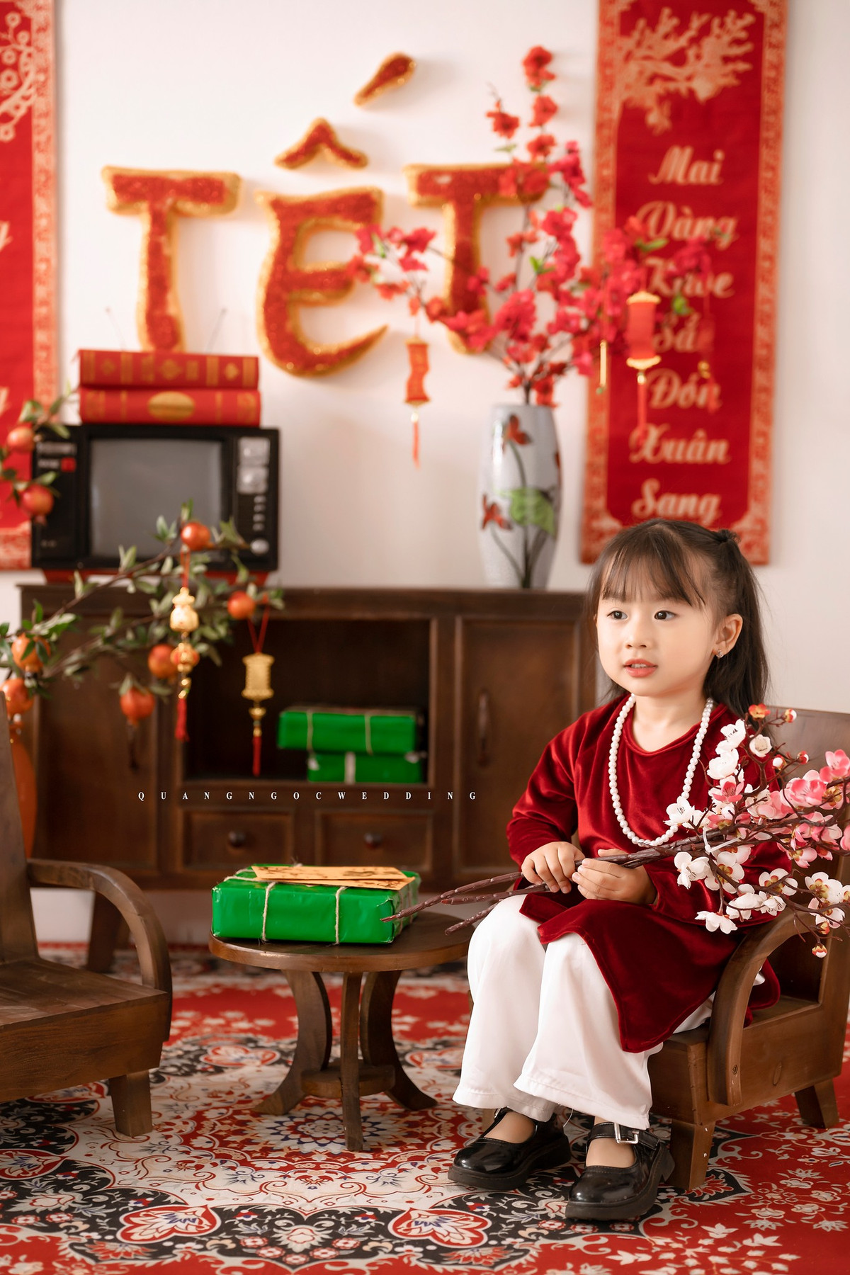 Em bé trong bộ ảnh diện áo dài nhung đỏ, cổ đeo chuỗi ngọc, đầu đội mấn ngồi trên ghế cổ tạo dáng rất hồn nhiên.