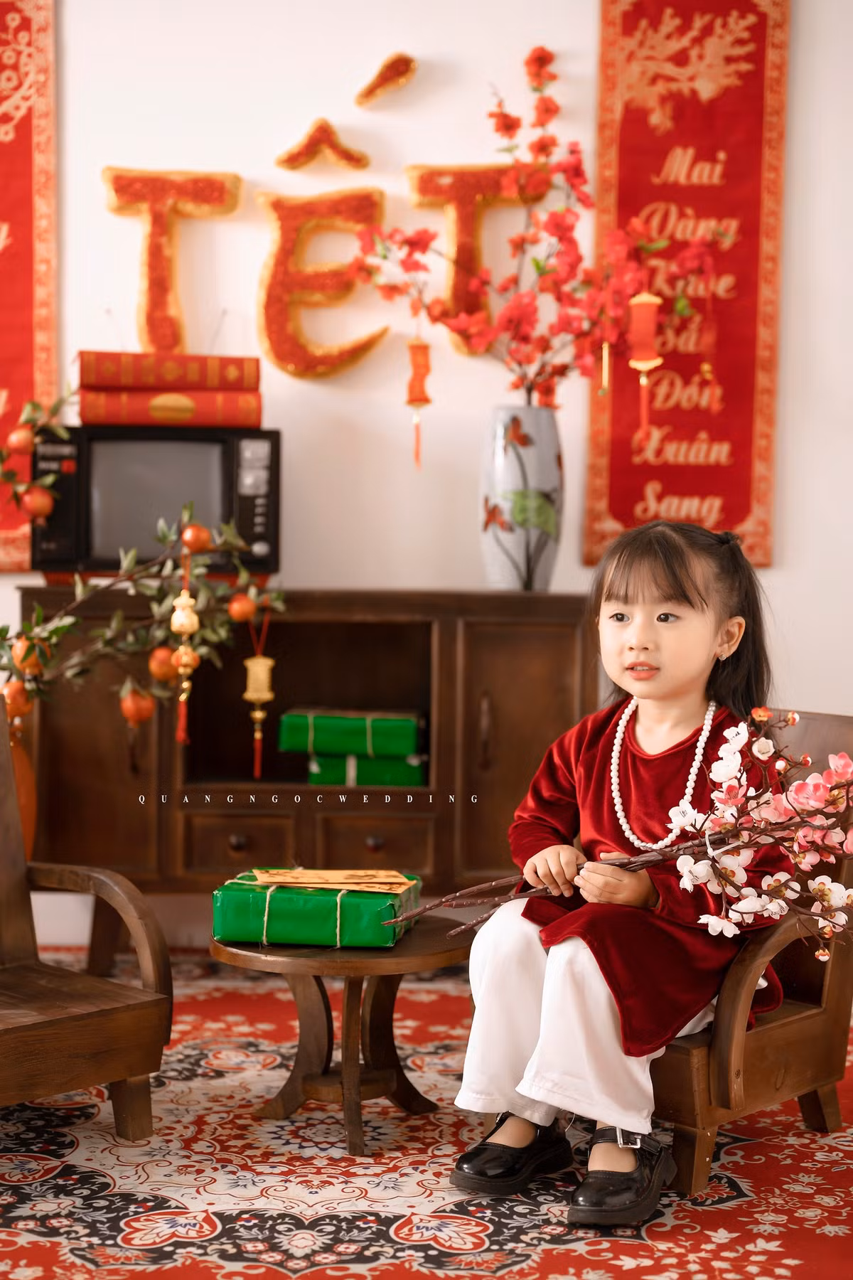 Em bé trong bộ ảnh diện áo dài nhung đỏ, cổ đeo chuỗi ngọc, đầu đội mấn ngồi trên ghế cổ tạo dáng rất hồn nhiên.