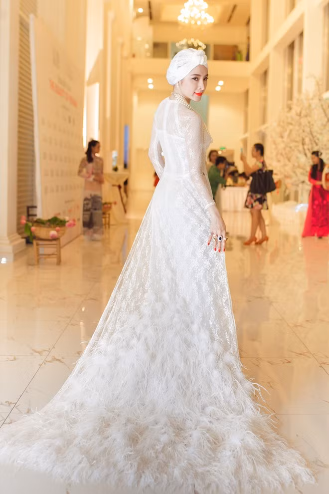 Áo dài chất liệu ren trắng của NTK Thuận Việt khá đặc biệt với tà sau dài, đính lông sang trọng. Kết hợp cùng bộ cánh lạ mắt, Angela Phương Trinh đội khăn turban ren cá tính. Người đẹp trang điểm đậm quyến rũ. Phụ kiện chuỗi hạt làm điểm nhấn phong cách thêm quý phái.