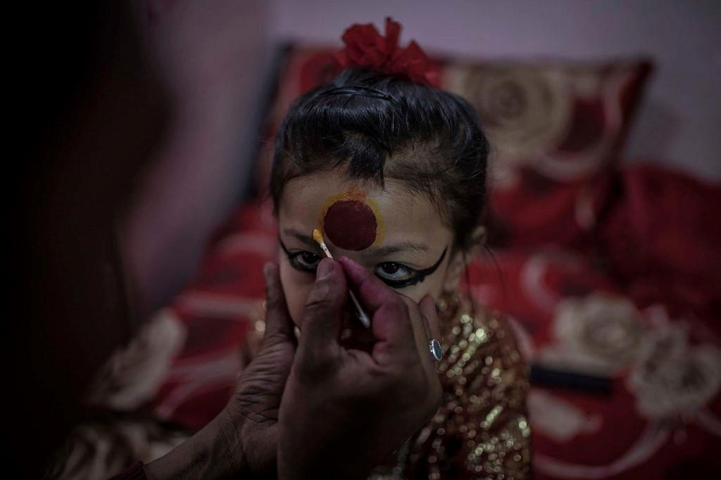 Không chỉ có một Kumari, nhưng những Kumari quan trọng nhất đến từ Patan và Kathmandu, theo Maria Contreras Coll, nhiếp ảnh gia đã tới Nepal năm 2017 và 2018 để gặp và chụp ảnh những cô gái đang và từng là Kumari. Bà cho biết nhiều lứa trẻ em gái đã được chọn làm Kumari từ ít nhất 300 năm nay. Trong ảnh, cha của nữ thần sống Patan đang trang điểm cho con gái mình trong đền Kumari ở Patan. Ảnh: Maria Contreras Coll.