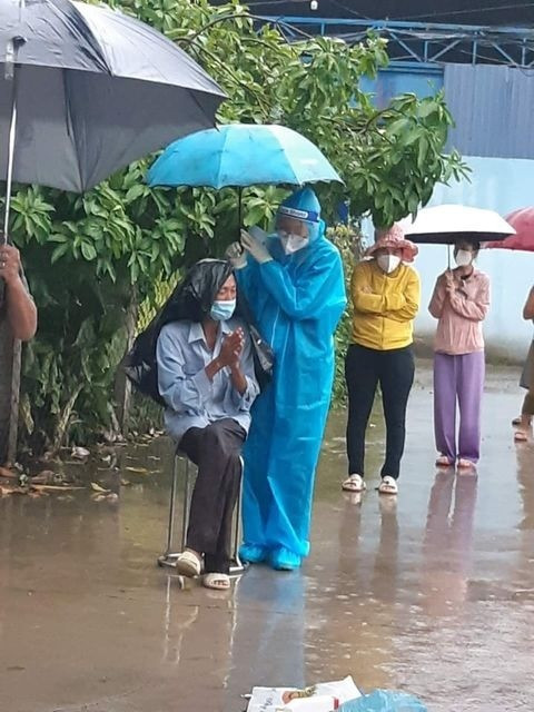 Hình ảnh tình nguyện viên che ô tránh mưa cho một người dân đang xếp hàng chờ.