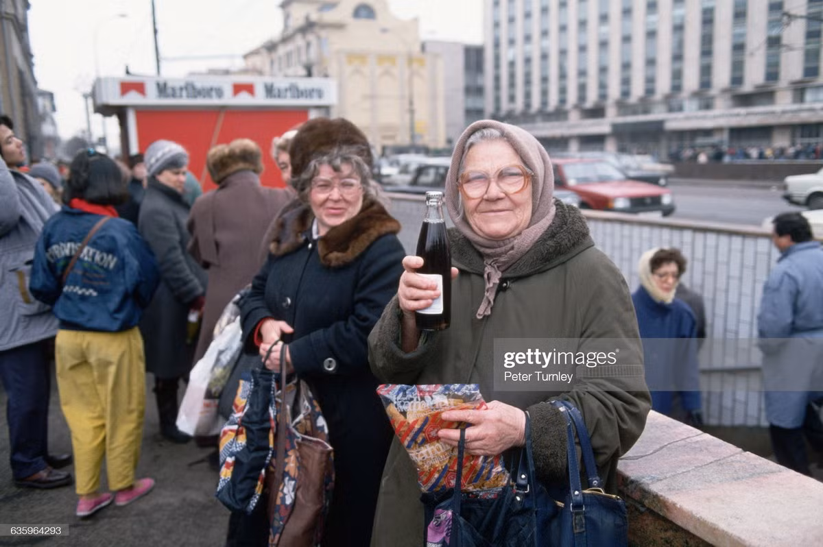 Cụ bà khoe chai Cola trên đường phố Moscow, ảnh chụp năm 1993. Phía xa là quầy thuốc lá Marlboro, một thương hiệu đến từ phương Tây.