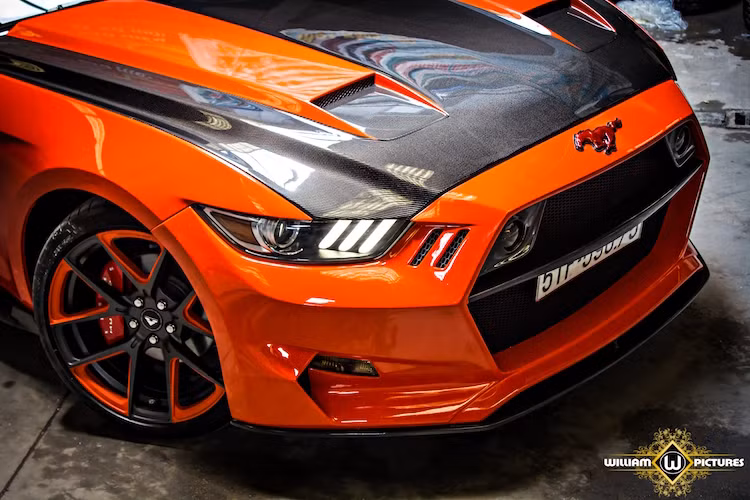 Đi kèm với bộ bodykit là nắp ca-pô bằng sợi carbon với khe hút gió làm mát mới, cộng với phần cản trước đã gần như hoàn toàn thay đổi thiết kế phần đầu của chiếc Mustang. Thay vì sơn hoàn toàn nắp ca-pô, chủ xe đã chỉ sơn màu cam trùng với thân xe dọc 2 khe hút gió và để trần lớp sợi carbon.