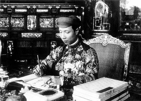 Chỉ có trên áo vua, hậu mới trang trí hoa văn tứ thời, bát bửu. Áo mão của vương tôn và cung giai, bát bửu được thay thế bằng những cổ đồ. Ảnh: Vua Khải Định trong trang phục thường triều, đầu đội khăn xếp, đang làm việc trong điện Cần Chánh.