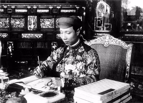 Chỉ có trên áo vua, hậu mới trang trí hoa văn tứ thời, bát bửu. Áo mão của vương tôn và cung giai, bát bửu được thay thế bằng những cổ đồ. Ảnh: Vua Khải Định trong trang phục thường triều, đầu đội khăn xếp, đang làm việc trong điện Cần Chánh.