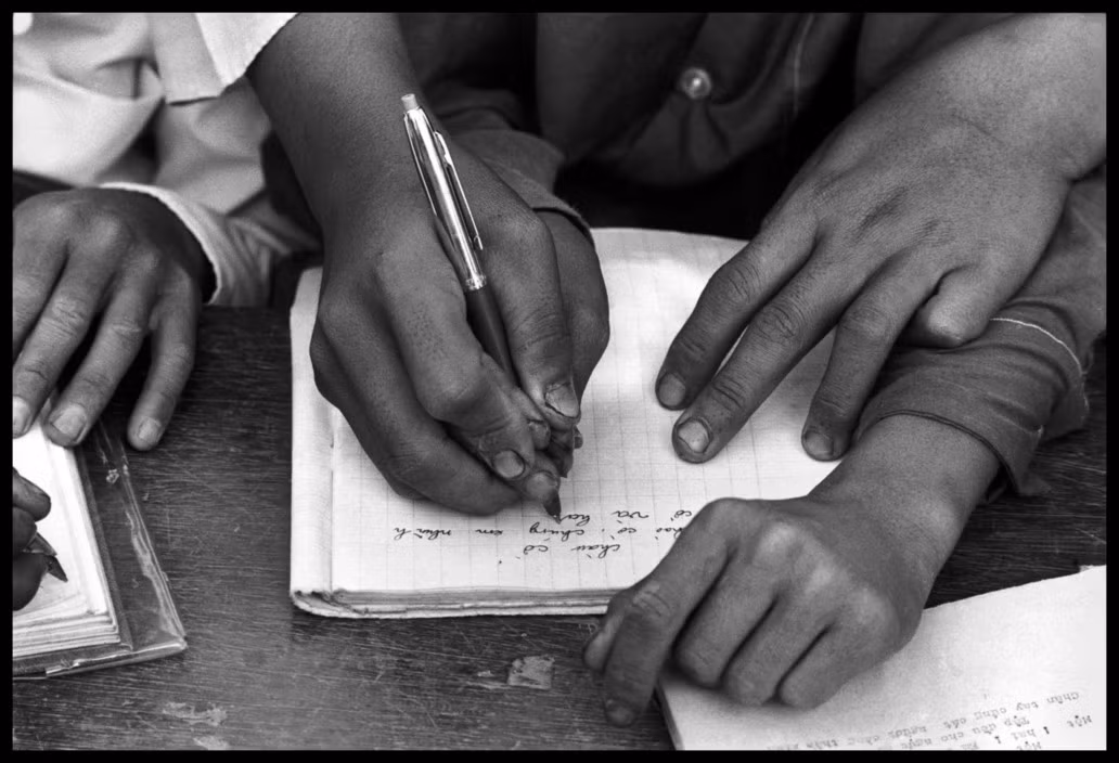 Giáo viên cầm tay học sinh để hướng dẫn cách viết chữ trog lớp học. Ảnh: Magnumphotos.com.