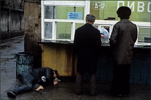 Hai người đàn ông đứng chờ mua bia ở một kios vào một hôm mưa gió trong khi một người đàn ông khác say rượu. Ảnh VT