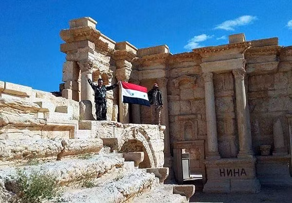 Hai binh sỹ Syria giơ cao cờ Syria tại một địa điểm lịch sử.