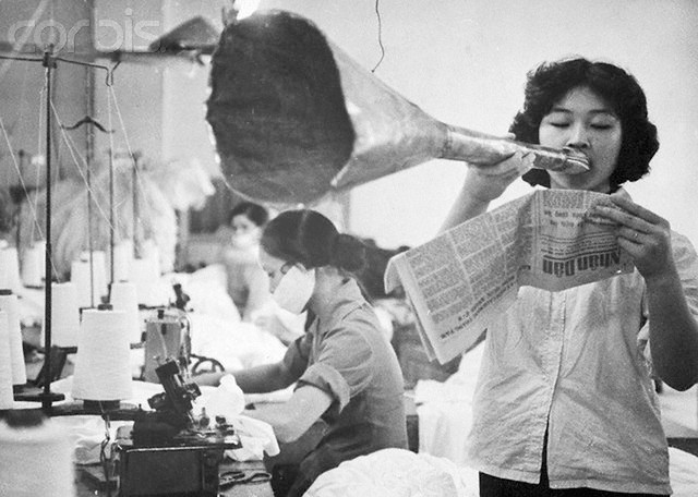Các nữ công nhân vừa làm việc vừa nghe Nguyen Thi Lien Khuong đọc những tin tức thời sự được đăng tải trên báo bằng một chiếc loa tự chế. Ảnh chụp tại một nhà máy may ở Hà Nội ngày 13/2/1968.