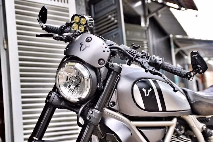 Khu vực xung quanh tay lái của Scrambler đã được chủ nhân thay đổi khá nhiều, trong đó thay vào vị trí của đồng hồ nguyên bản là một đèn LED chiếu sáng tăng cường, trong khi cụm đồng hồ nguyên bản được chuyển xuống chảng 3 gọn gàng hơn. Ghi-đông nguyên bản của Scrambler đã được thay bằng cặp tay lái clip-on thể thao hơn, nằm phía trên là cùm côn Accossato và cùm phanh Brembo RCS 19.