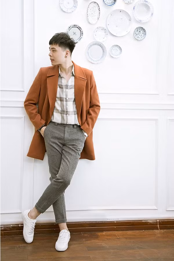 Nam ca sĩ phối áo sơ mi kẻ với quần vải xám và áo khoác dáng dài rất lịch lãm, trẻ trung.