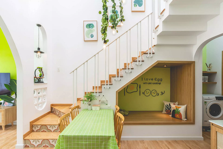 Khu bếp xinh xắn được bố trí thêm một tủ thấp, tăng không gian lưu trữ. Những hình vẽ tay trang trí, tạo điểm nhấn độc đáo cho bếp. Cầu thang của công trình nằm trong khu bếp kết hợp với bàn ăn, tận dụng không gian dưới cầu thang để bố trí thêm một góc thư giãn khi nấu và chờ đồ ăn.