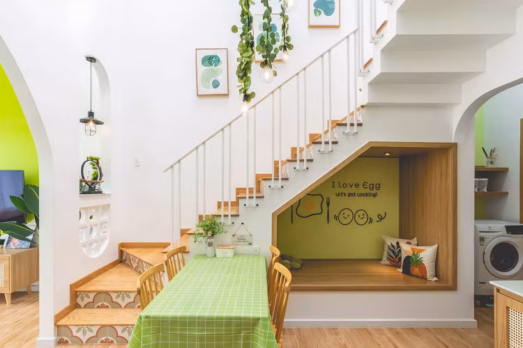 Khu bếp xinh xắn được bố trí thêm một tủ thấp, tăng không gian lưu trữ. Những hình vẽ tay trang trí, tạo điểm nhấn độc đáo cho bếp. Cầu thang của công trình nằm trong khu bếp kết hợp với bàn ăn, tận dụng không gian dưới cầu thang để bố trí thêm một góc thư giãn khi nấu và chờ đồ ăn.