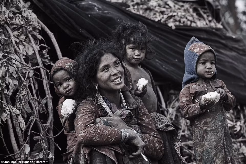 Một phụ nữ trong bộ tộc Raute ngồi hút thuốc cạnh 3 đứa con nhỏ bên ngoài túp lều của họ.