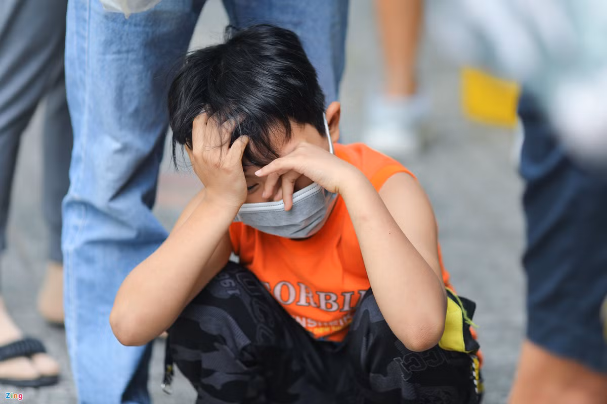 Bé Anh Hào (10 tuổi) ngồi bệt trong lúc xếp hàng chờ. Phụ huynh bé Hào cho biết một ngày trước, bé có biểu hiện sốt cao nhưng test nhanh tại nhà vẫn âm tính. Đến sáng nay, gia đình đưa bé lên bệnh viện xét nghiệm thì kết quả dương tính. Do lượng trẻ tại bệnh viện quá đông, 2 cha con phải chờ gần 2 tiếng mà chưa tới lượt khám sàng lọc.