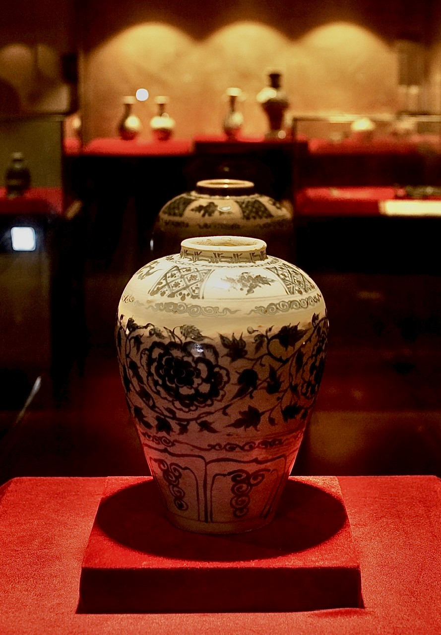 Chum gốm men trắng vẽ lam, trang trí hoa cúc mãn khai. Vai có gấm hình trám vẽ hoa lá. Niên đại thế kỷ 15 của nhà sưu tầm Vũ Anh Tuấn.