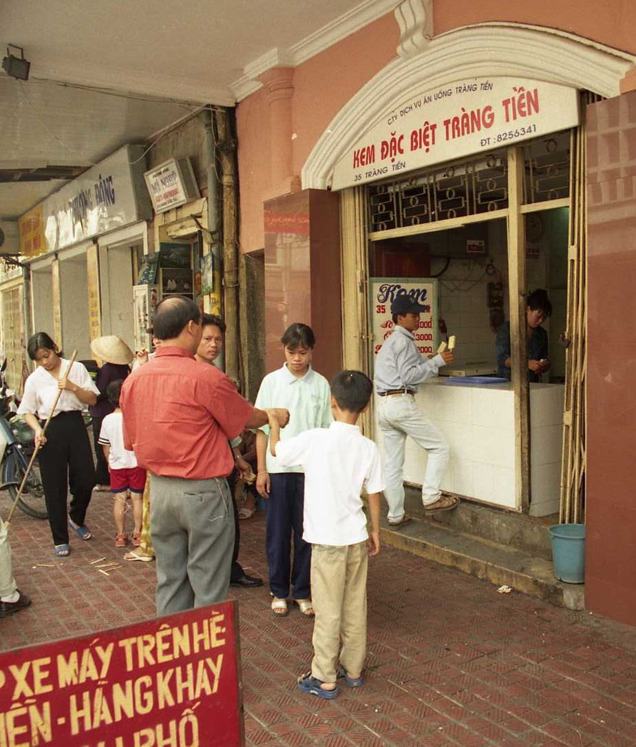 Một nữ nhân viên quét dọn những que kem khách hàng xả xuống vỉa hè sau khi ăn. Trước cửa hàng có tấm biển hướng dẫn chỗ để xe máy. Ảnh chụp năm 1998. Ảnh: Yuichi Kobayashi.