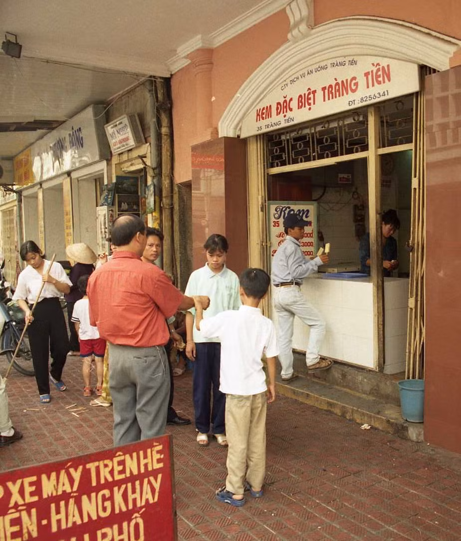Một nữ nhân viên quét dọn những que kem khách hàng xả xuống vỉa hè sau khi ăn. Trước cửa hàng có tấm biển hướng dẫn chỗ để xe máy. Ảnh chụp năm 1998. Ảnh: Yuichi Kobayashi.