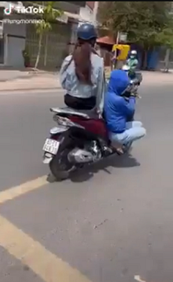 Một cô gái mặc áo chống nắng kín mít, điều khiển chiếc xe máy SH đang chở phía sau 1 cô gái khác. Cô gái điều khiển xe máy vừa đi vừa ngồi bệt xuống chỗ để chân, 1 chân duỗi thẳng, quệt xuống đường.