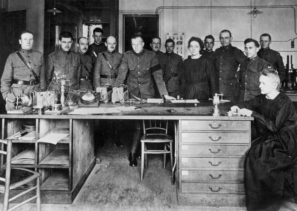 Giáo sư Marie Curie (phải) đang làm việc tại một phòng thí nghiệm ở trường đại học Paris. Xung quanh là các học sinh của bà gồm các nhà vật lý học trẻ và các thành viên của lực lượng công tác Mỹ.