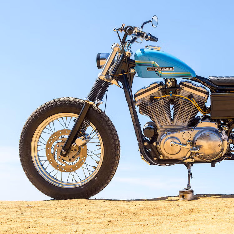 Sau khi đã lột hết các chi tiết, chiếc 883 chỉ còn giữ lại khung và máy. Clint đã bắt đầu lắp cặp lốp gai Firestone ANS vào vành và chỉnh dáng. Phuộc trước có lò xo mềm, chịu lực cao hơn và vỏ phuộc sơn màu đen.