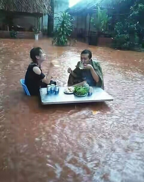 Bất chấp nước lũ lênh láng khắp sân, 2 thanh niên vẫn vô tư nâng cốc bia và nhâm nhi mồi.