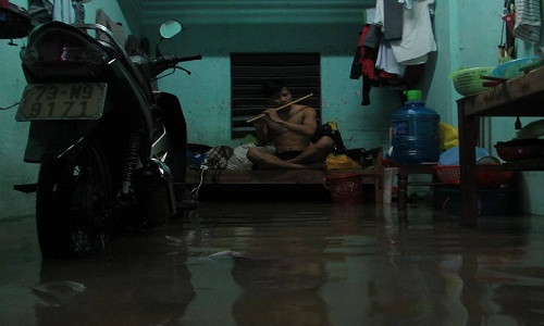 Một nam sinh viên ngồi trên giường thổi sáo trong lúc nước lụt "tấn công" vào phòng trọ.