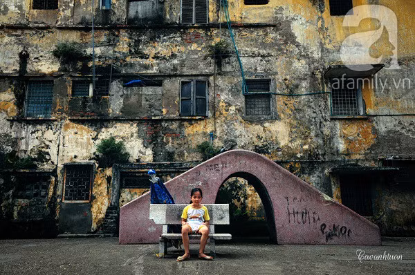 Một em bé ngồi trên ghế đá ở khu vui chơi của trẻ em, phía sau là bức tường loang lổ đầy dấu vết của thời gian.