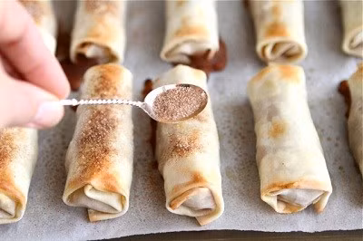 Đánh tan trứng trong 1 chén nhỏ. Sau đó, trải từng lá bánh cuốn lên thớt và dùng cọ quét trứng dọc theo các cạnh. Dùng thìa xúc khoảng 30g nhân táo đặt vào lá bánh. Tiếp đó, lần lượt gấp các cạnh lại rồi khéo léo cuốn thật tròn và chặt tay. Sau đó, bạn đặt bánh táo vào khay nướng đã được lót sẵn giấy nến. Ảnh: monngon.asia.vn.