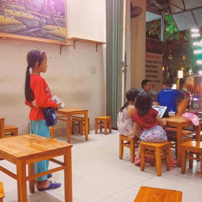 Một bé gái cầm trên tay xấp vé số, đi dép lê mải nhìn theo hai đứa trẻ khác ăn mặc chỉn chu ngồi chơi máy tính bảng. Ánh mắt cô bé nghèo chằm chằm dõi theo khiến người xem không khỏi chạnh lòng. Ảnh: Trương Minh Hiếu.