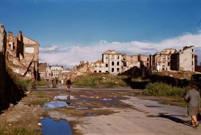 Một vài người dân đi lại trên con đường với hai bên là những tòa nhà, công trình hư hại do trúng bom đạn của phát xít Đức.