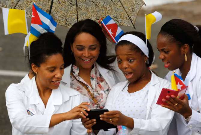 Các y tá vừa cầm cờ Vatican và Cuba vừa xem điện thoại trong lúc chờ xe của Giáo hoàng đi qua.