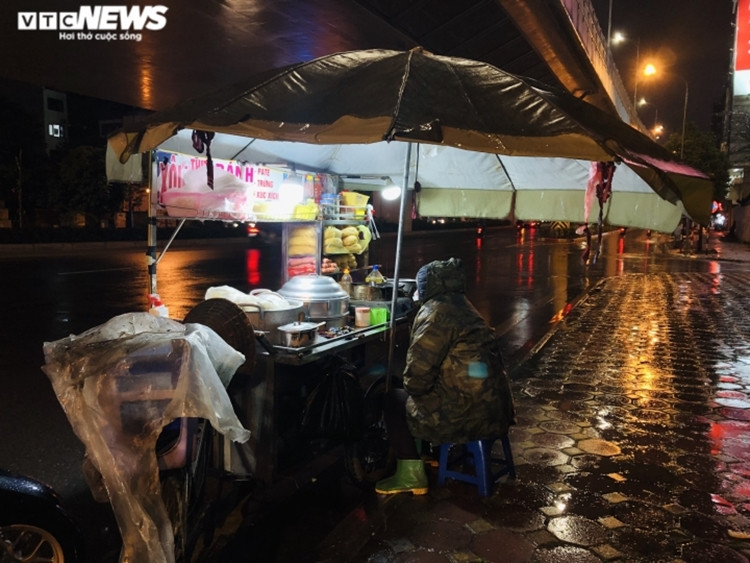 Bà Nguyễn Thị Hà (55 tuổi) ngồi co ro bên chiếc xe đẩy bán xôi, ngô, bánh mì. Trời đã về khuya, nhưng bà vẫn cố chờ thêm vài phút với hi vọng sẽ có khách mua ủng hộ được đồng nào hay đồng đó. "Tối giờ tôi mới bán được 3 bắp ngô và 1 cái bánh mì. Mưa lạnh như này ít người ra đường lắm, chỉ có lao động nghèo như tôi mới phải cày cuốc thôi", bà Hà buồn bã nói. (Ảnh: VTC News).