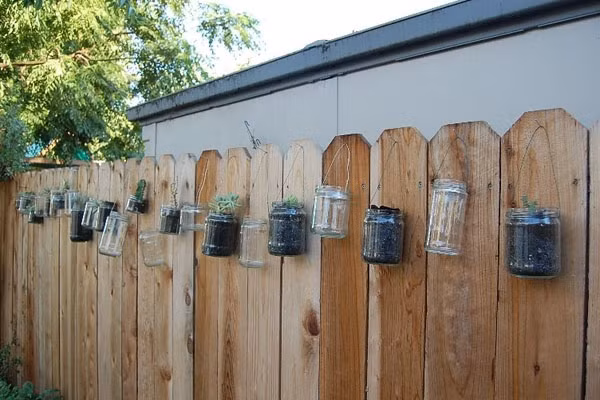 Tận dụng lọ thủy tinh Những chiếc lọ thủy tinh cũng có thể được tận dụng một cách sáng tạo để làm đẹp cho hàng rào. Sau khi sử dụng thực phẩm bên trong, bạn bóc nhãn mác, làm sạch lọ và bỏ đất bên trong. Hãy so le một lọ trồng cây một lọ để không và treo những chiếc lọ thẳng hàng trên hàng rào. Mỗi khi đến dịp lễ, Tết, bạn có thể bỏ nến hay đèn LED bên trong những hộp thủy tinh để trống, giúp hàng rào đẹp lung linh và lãng mạn khi đêm về.