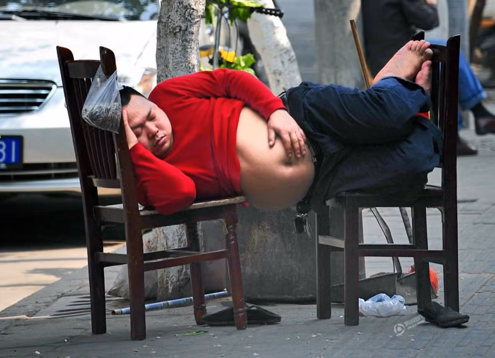 Người đàn ông béo nằm ngủ ngay trên vỉa hè với hai chiếc ghế. (Nguồn: QQ)