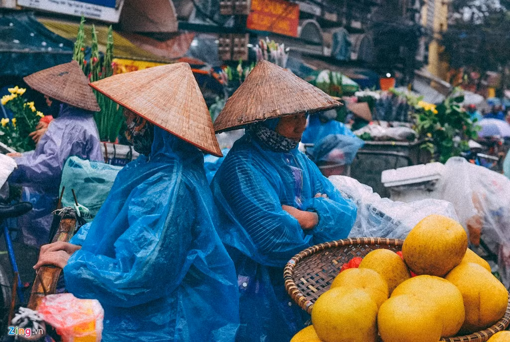 Nhóm phụ nữ bán hàng trước chợ Cầu Đông đứng nhìn dòng người qua lại. Trong những ngày mưa, việc đứng bán hàng ngoài trời vất vả hơn nhiều so với những ngày tạnh ráo, còn thu nhập lại bị giảm do lượng khách mua hàng thưa thớt hơn.